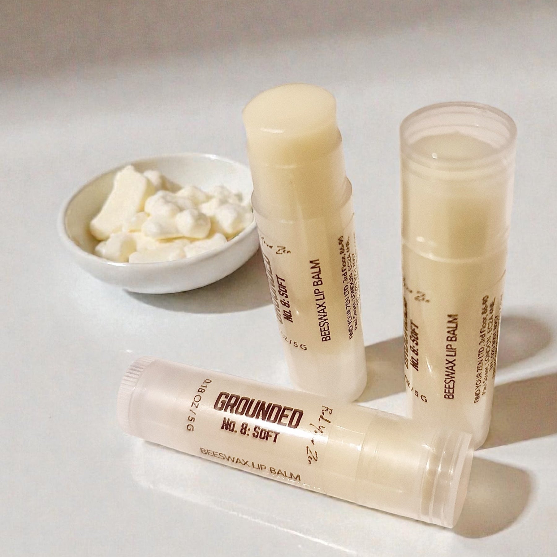 Three lip balms on a white surface with a small bowl of mango butter in the background.